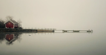 Ein kleines rotes Haus steht am Ufer eines stillen Sees reflektiert im Wasser neblige Atmosphäre und ein langer Steg erstreckt sich übers Wasser.