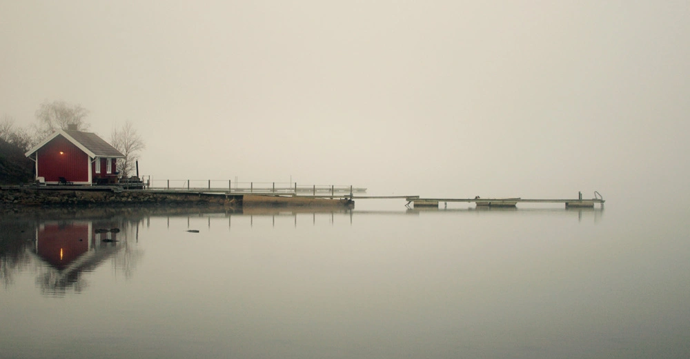 Ein kleines rotes Haus steht am Ufer eines stillen Sees reflektiert im Wasser neblige Atmosphäre und ein langer Steg erstreckt sich übers Wasser.