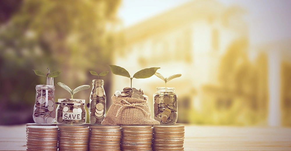 Münzbehälter und ein Säckchen voller Münzen mit wachsenden Pflanzen darauf stehen auf stapelweise Münzen in einem unscharfen natürlichen Hintergrund mit einem Gebäude. Ein Glas ist mit dem Wort SAVE beschriftet.