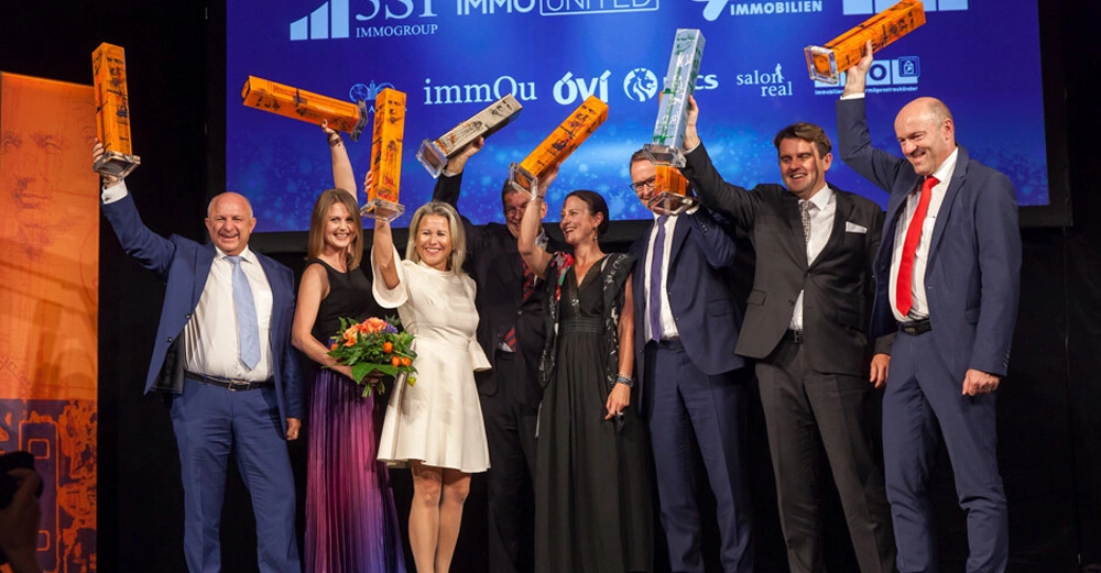 Mehrere Personen stehen auf einer Bühne und halten große orangefarbene und durchsichtige Trophäen in die Luft sie tragen formelle Kleidung und lächeln; im Hintergrund befinden sich Logos verschiedener Unternehmen.