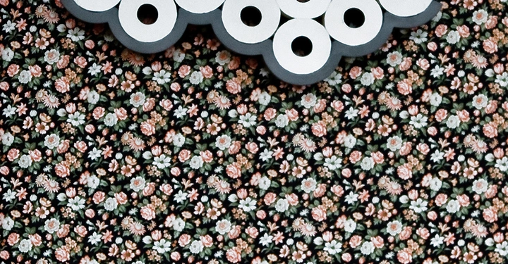 Toilettenpapierrollen hängen in einer Wolkenform an der Wand über einer weißen Toilette vor einer blumig gemusterten Tapete im Badezimmer