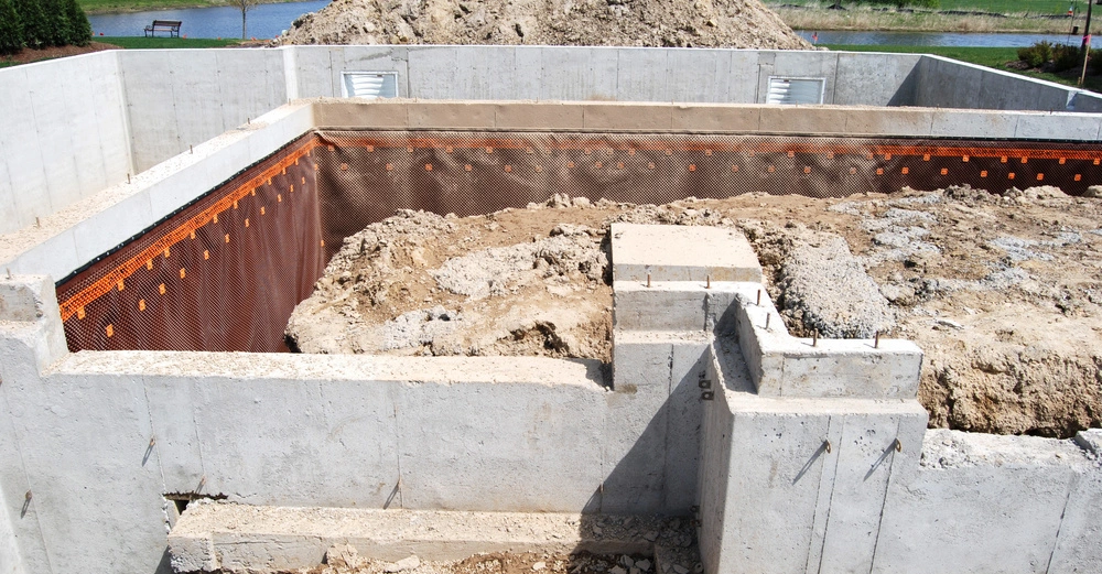 Betonfundament wird gebaut und ist teilweise ausgegraben Umgebung zeigt Erdberge und Teiche mit Häusern im Hintergrund.