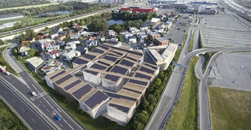 Komplex mit vielen Gebäuden ist umgeben von Straßen und kleineren Häusern in einer städtischen Umgebung. Links führt eine Autobahn, rechts ist ein großer leerer Parkplatz zu sehen. Text: B17 PASN450.