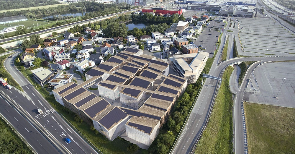 Komplex mit vielen Gebäuden ist umgeben von Straßen und kleineren Häusern in einer städtischen Umgebung. Links führt eine Autobahn, rechts ist ein großer leerer Parkplatz zu sehen. Text: B17 PASN450.