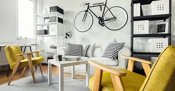 Ein schwarzes Fahrrad hängt an der Wand oberhalb eines weißen Sofas mit gemusterten Kissen in einem modernen Wohnzimmer mit gelben Stühlen Regalen und einem weißen Tisch.