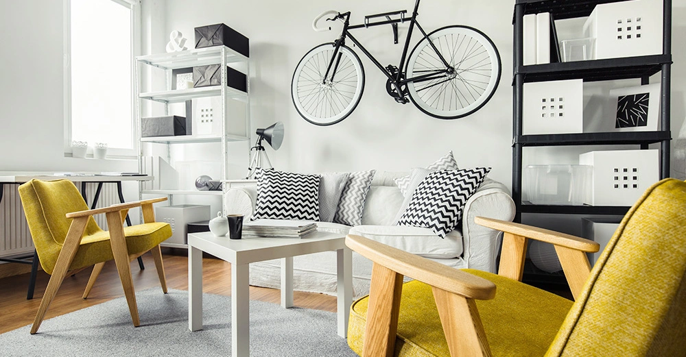 Ein schwarzes Fahrrad hängt an der Wand oberhalb eines weißen Sofas mit gemusterten Kissen in einem modernen Wohnzimmer mit gelben Stühlen Regalen und einem weißen Tisch.