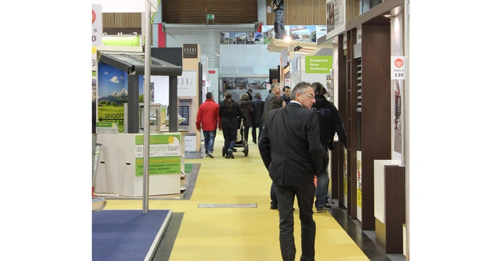 Galerie-Bild 17: Menschen gehen in einer Messehalle auf einem gelben Weg zwischen verschiedenen Messeständen spazieren. Schilder und Plakate mit Text wie "die pergarten-bauer", "Halle 6 130" und "Behaglichkeit Wärme Wohlbefinden" sind sichtbar.