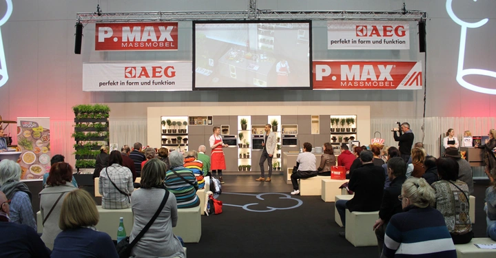 Galerie-Bild 17: Mehrere Personen hören einer Präsentation in einer Ausstellungshalle zu. Zwei Referenten sprechen auf der Bühne vor einer Küchenzeile. Banner oben zeigen P.MAX MASSMÖBEL und AEG perfekt in Form und Funktion.