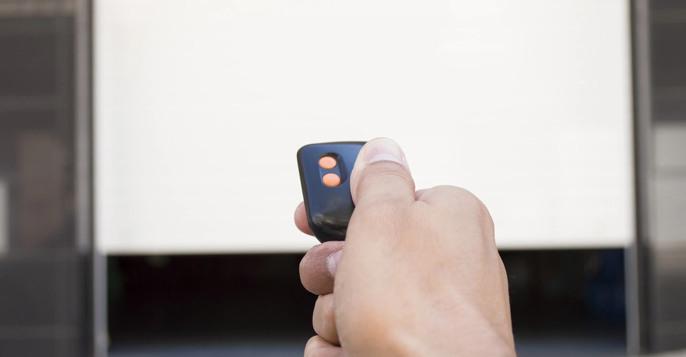 Hand hält eine Fernbedienung vor einem teilweise geöffneten Garagentor in einem modernen Stadtumfeld.