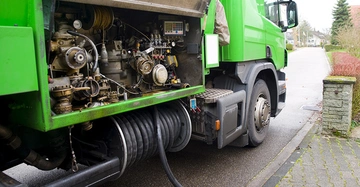grüner Öltank-Laster auf Straße in Wohngebiet, der über große Leitung Heizöl in Heizkeller leitet