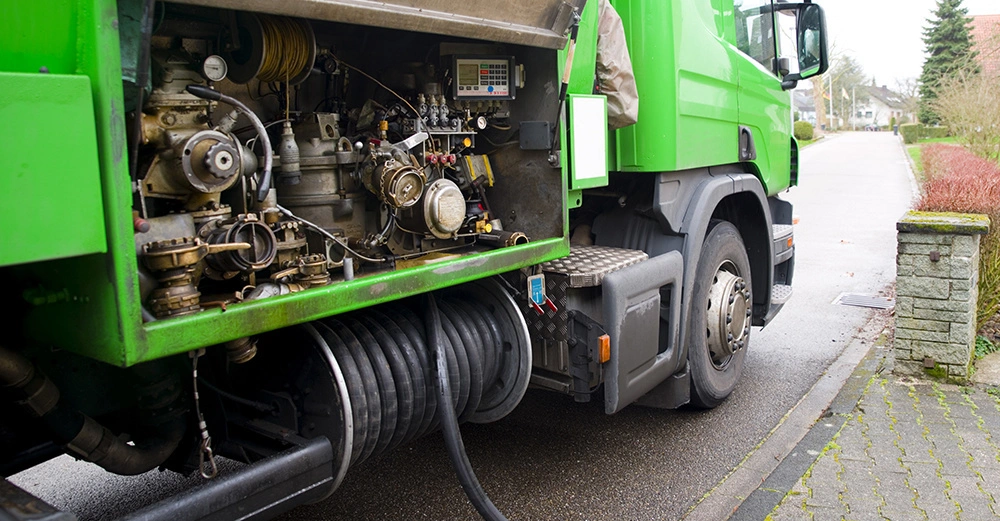 grüner Öltank-Laster auf Straße in Wohngebiet, der über große Leitung Heizöl in Heizkeller leitet