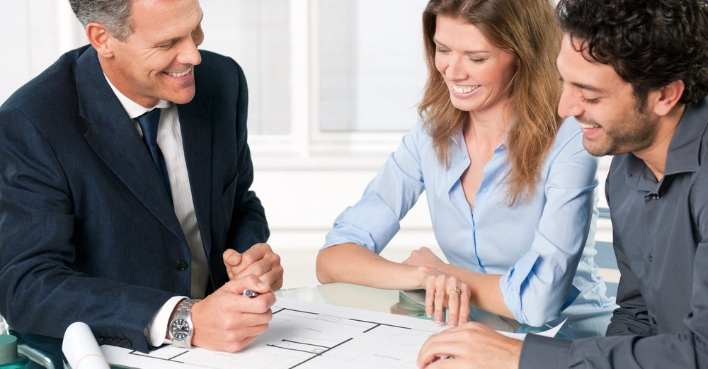 Ein Geschäftsmann erklärt einem Paar Baupläne auf einem Tisch in einem Büro. Alle drei Personen sind lächelnd und konzentriert auf die Zeichnungen vor ihnen.