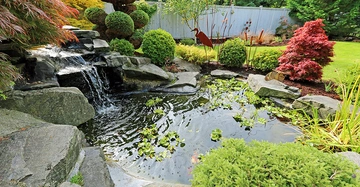 Ein kleiner Teich mit Seerosen reflektiert eine Wasserfall-Rock-Struktur die ihn speist umgeben von üppigen Sträuchern Bäumen und dekorativen Tierfiguren in einem gepflegten Garten mit Rasen und Holzzaun im Hintergrund
