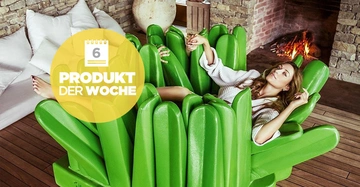 Eine Frau liegt entspannt in einem großen grünen Stuhlsessel im Kaktus-Design in einem gemütlichen Wohnzimmer mit Kamin. Text: 6 PRODUKT DER WOCHE