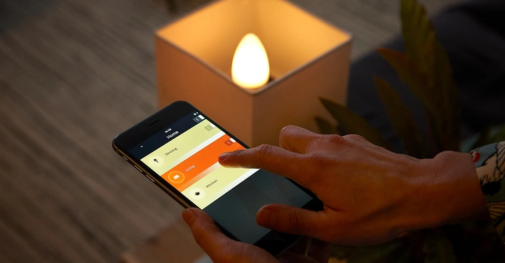 Eine Hand hält ein Smartphone und steuert eine Beleuchtungs-App Wohnzimmer im Hintergrund ein warmer heller Lampenschirm auf einem Tisch in einem gemütlichen Wohnraum. Text Home Dimmer Living Kitchen