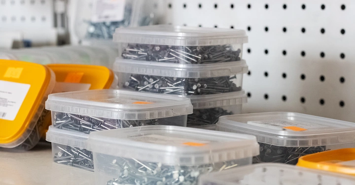 mehrere Kunststoffbehälter mit gelbem Deckel, voll mit Nägeln unterschiedlicher Größe, gestapelt in einem Baumarkt-Regal