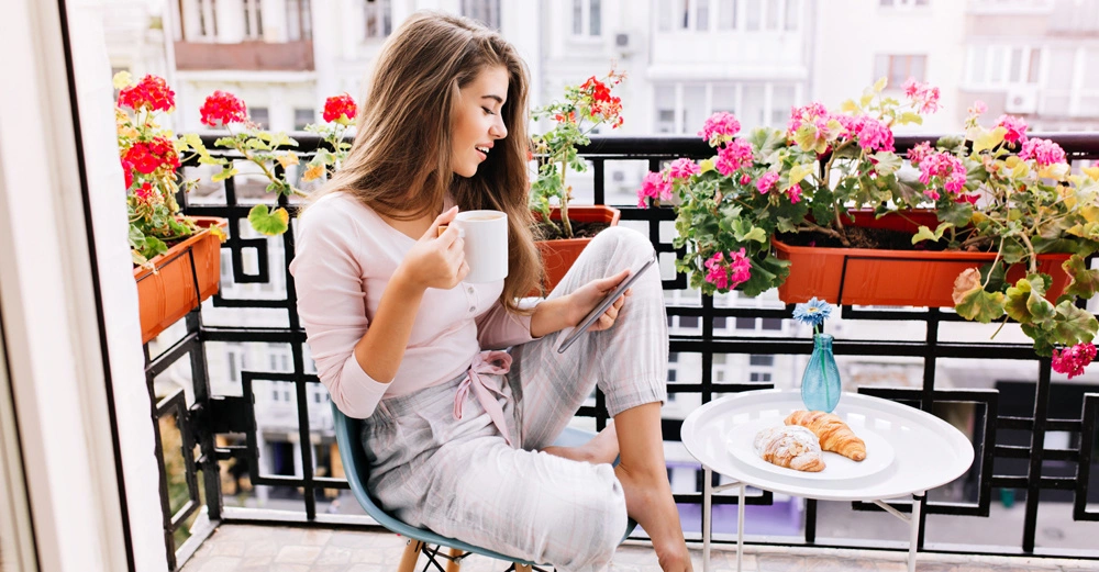 Eine Frau sitzt auf einem Balkon trinkt Kaffee und benutzt ihr Smartphone Die Umgebung ist mit blühenden Blumen geschmückt und auf einem kleinen Tisch stehen Teller mit Gebäck und eine Vase