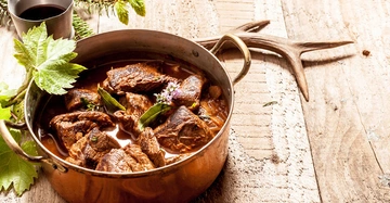 Kupfertopf mit geschmortem Fleisch und Kräutern stehend auf einem rustikalen Holztisch im Freien neben einem Geweih und einem Metallbecher mit dunklem Getränk umgeben von Tannenzweigen und grünen Blättern