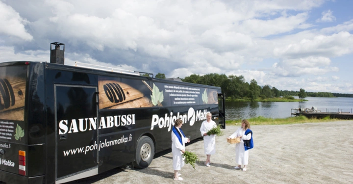 Ein schwarzer Bus beschriftet mit SAUNABUSSI und Pohjolan Matka steht auf einem Schotterplatz. Drei Personen in weißen Bademänteln stehen daneben und halten Pflanzen und einen Korb. Im Hintergrund befindet sich ein See mit einem Holzsteg und umgeben von Bäumen.