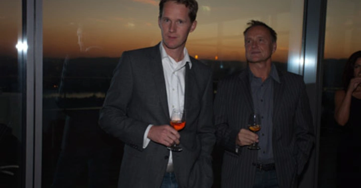 Galerie-Bild 66: Zwei Männer halten Getränke während sie in einem modernen Raum mit großen Fenstern stehen Die Abenddämmerung ist draußen sichtbar Im Hintergrund sind schemenhaft weitere Personen zu erkennen