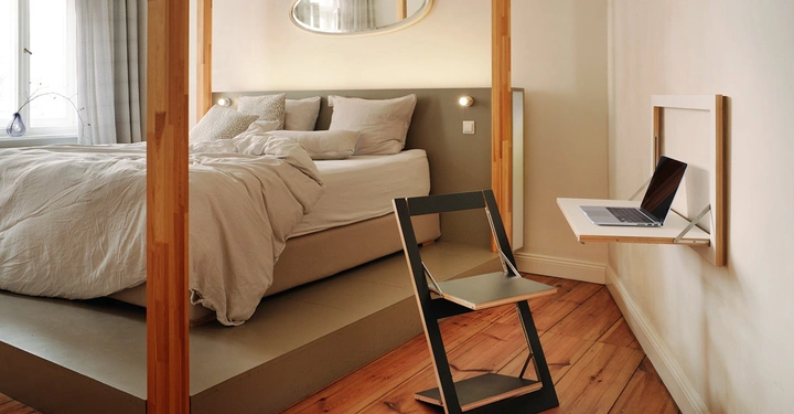 Ein gemütliches Schlafzimmer mit einem großen, gepolsterten Bett, einem Holzstuhl und einem an der Wand montierten Klapptisch, auf dem ein Laptop steht; helle Wände und Holzboden.