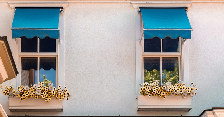 Hausfassade mit zwei kleinen Kastenfenstern, darüber tieflbaue, mediterran wirkende Fenstermarkisen. Blumen in Trögen hängen vor den Fenstern. Sonniges Ambiente