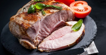 Ein saftiges Stück gebratenes Schweinefleisch liegt auf einem schwarzen Schieferbrett mit frischen Basilikumblättern und Tomaten garniert im Hintergrund. Ein Messer und eine Gabel liegen daneben auf einer gestreiften Serviette.