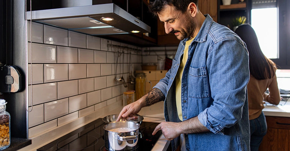 Mann rührt Essen im Kochtopf auf Induktionsherd in moderner Küche, Frau wäscht Geschirr im Hintergrund.