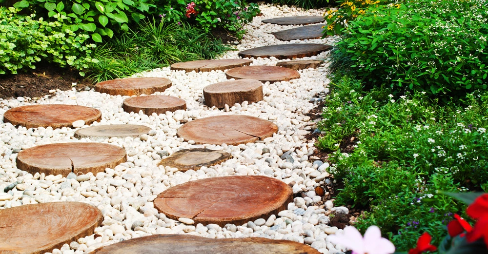 Holzscheiben bilden einen geschwungenen Pfad auf weißen Kieseln. Sie führen durch einen üppig grünen Garten mit blühenden Pflanzen und bunten Blumen.