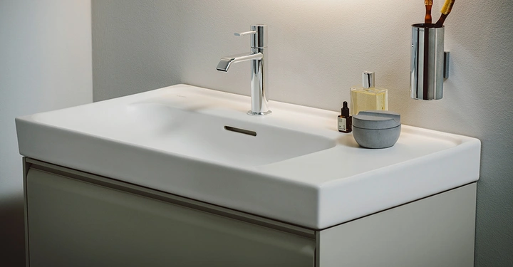 Ein weißes Waschbecken mit einer verchromten Armatur steht in einem modernen Badezimmer. Auf dem Waschbecken befinden sich ein Parfümfläschchen ein kleiner Behälter und eine Flasche. An der Wand hängt ein Zahnbürstenhalter.