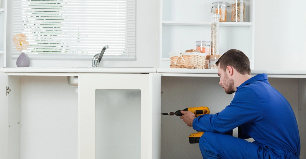 Arbeiter in blauer Uniform bohrt ein Loch in einen weißen Küchenschrank in einer modernen Küche mit weißen Schränken und Küchenutensilien im Hintergrund.