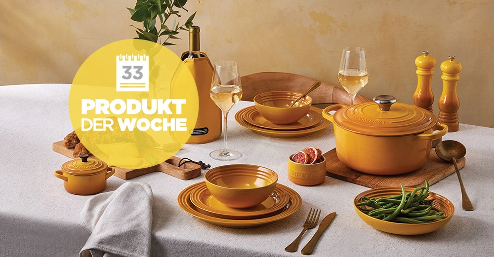 Gelbes Geschirr steht dekorativ auf einem gedeckten Tisch mit weißer Tischdecke. Zwei Gläser Weißwein, Besteck, grüne Bohnen und ein Handtuch sind ebenfalls zu sehen. In einem gelben Kreis steht "33 PRODUKT DER WOCHE".