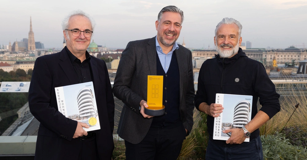 Drei Männer halten Auszeichnungen auf einer Dachterrasse mit Blick auf eine Stadt im Hintergrund. Der mittlere Mann hält eine gelbe Trophäe mit der Aufschrift: Award Wohnbauten des Jahres 2023 Acknowledgement Quadriga Hochschule.