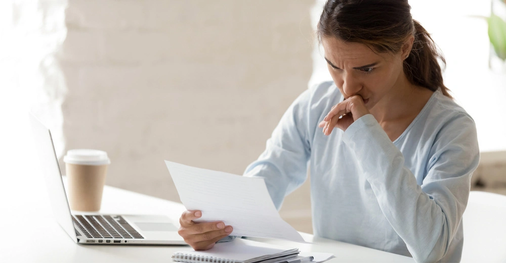 Junge Frau hält sitzt am Schreibtisch und blickt stirnrunzelnd auf ein Blatt Papier. Am Tisch neben ihr stehen ein Laptop und ein Einweg-Kaffeebecher.
