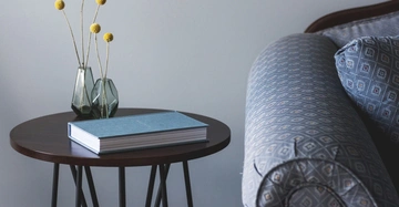 Buch liegt geschlossen auf rundem Holztisch mit zwei kleinen Vasen und gelben Blumen neben geschmücktem Sofa in einem Wohnzimmer.