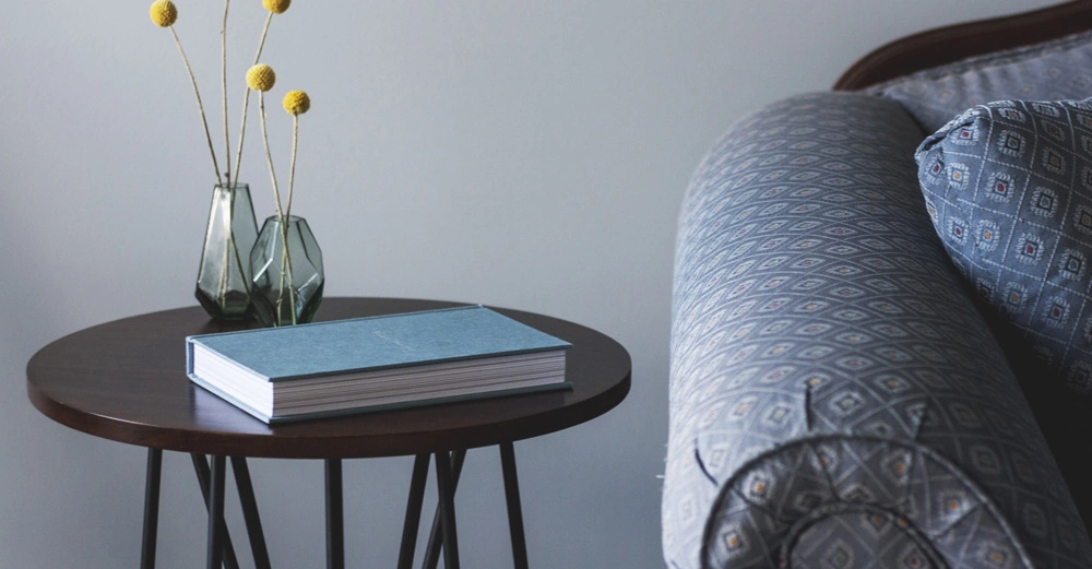 Buch liegt geschlossen auf rundem Holztisch mit zwei kleinen Vasen und gelben Blumen neben geschmücktem Sofa in einem Wohnzimmer.