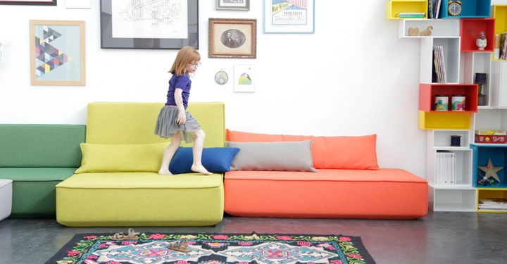 Ein Kind geht auf hellen Sofas in einem modernen Wohnzimmer mit bunten Regalen, Bildern an der Wand und einem bunten Teppich auf dem Boden.