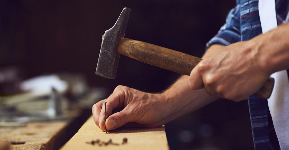 Mann in weißer Arbeitslatzhose und blau kariertem Hemd schlägt Nagel in Holzbrett ein