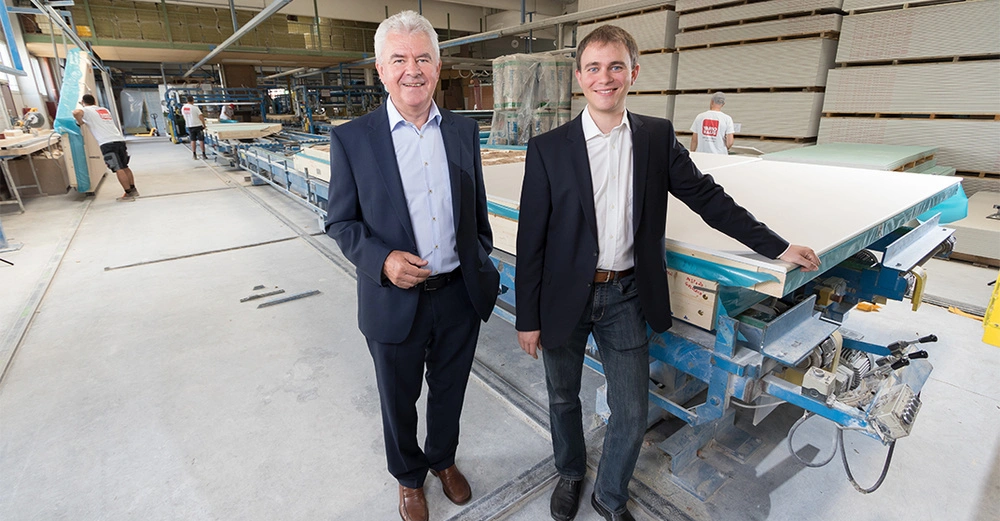 Zwei Männer in Anzügen stehen in einer industriellen Produktionshalle mit Maschinen und arbeiten; im Hintergrund arbeiten zwei Personen an Holzplatten.