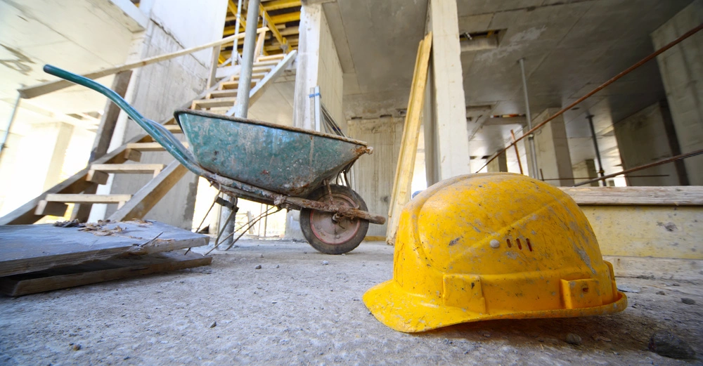 Ein gelber Bauhelm liegt neben einer Schubkarre auf Betonboden in einem unfertigen Gebäude mit Betonstützen und Treppen im Hintergrund.