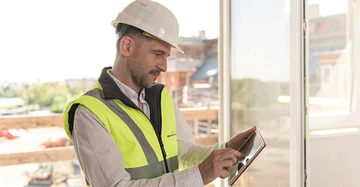 Bauarbeiter benutzt ein Tablet in einem Gebäude am offenen Fenster eine Baustelle ist im Freien sichtbar trägt gelbe Warnweste und weißen Schutzhelm.