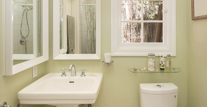 Waschbecken mit Spiegeln hängt an einer hellgrün gestrichenen Badezimmerwand daneben ein Fenster mit Blick auf Bäume Toilettenschüssel unter Regal mit Toilettenartikeln Dusche im Hintergrund links