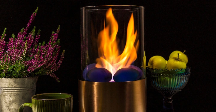 Kupferfarbene Tischlaterne mit aufflammendem Feuer auf zylindrischer Basis steht zwischen einem Metalltopf mit violetten Blumen links und einer grünen Schale mit Äpfeln rechts; grüne Tasse vorne links.