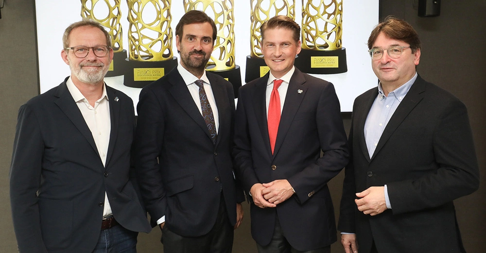 Hannes Horvath, Georg Flödl, Matthias Gass und Anton Holzapfel stehen nebeneinander und lächeln in die Kamera. Alle tragen einen dunklen Anzug. Auf einem Bildschirm im Hintergrund ist die goldene Trophäe des Prix d'Excellence zu sehen. 