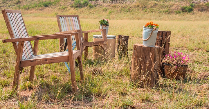 zwei Holzlehnsessel stehen auf trockenem, gemähtem Feld, Zwei Holzstämme als Tisch stehen daneben, auf einem ein Blechkübel mit schönen Blumen. Es wirkt sommerlich heiß