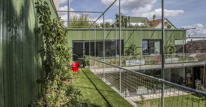Galerie-Bild 4: Das grüne Haus hat eine Terrasse mit Pflanzen links sitzt ein roter Stuhl das Gebäude hat große Fenster und befindet sich in einem grünen Garten bei sonnigem Wetter