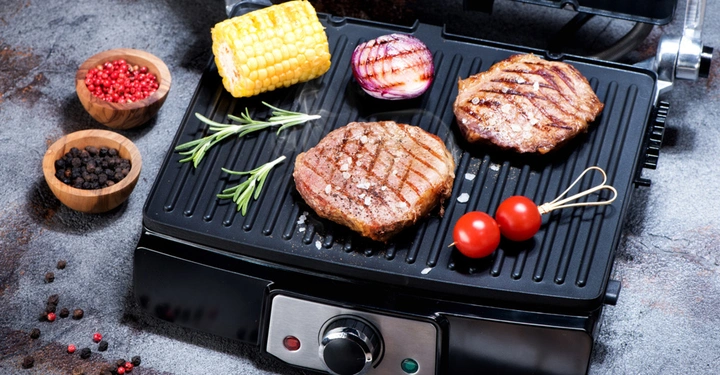 Zwei gegrillte Steaks grillen auf einem Elektrogrill daneben Maiskolben Tomaten und eine Zwiebel im Hintergrund Schalen mit Pfeffer und Gewürzen auf einer grauen Oberfläche