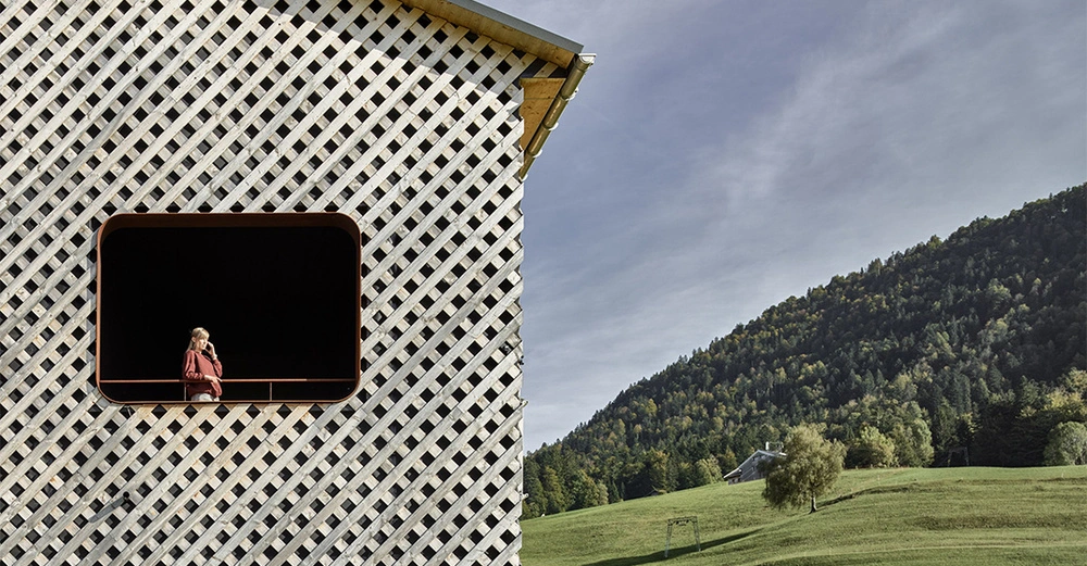 Person eine Frau schaut aus einem Fenster mit Holzlattenmuster Haus in ländlicher Gegend bewaldete Hügel und Himmel im Hintergrund