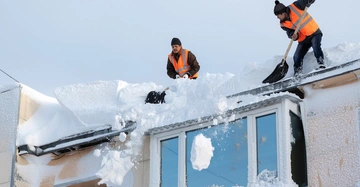 zwei Männer in orangen Warnwesten schaufeln mit Schneeschaufeln meterhohe Schneemassen von einem Dach