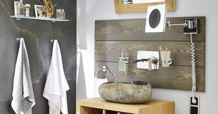 Galerie-Bild 6: Waschbecken-Set mit Steinschale steht auf Holzregal gefüllt mit Toilettenartikeln umgeben von Handtüchern und Spiegel in modernem Badezimmer mit dunklen Wandfliesen und Glasregal.